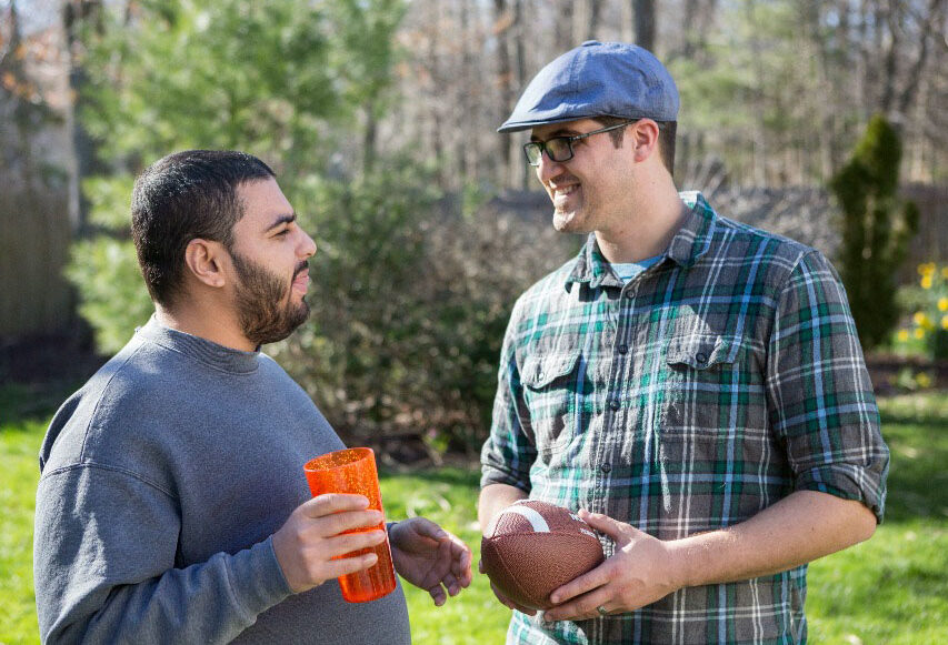 Two men talking in a forested area. Man on the left holding an orange cup and man on the right holding a football.